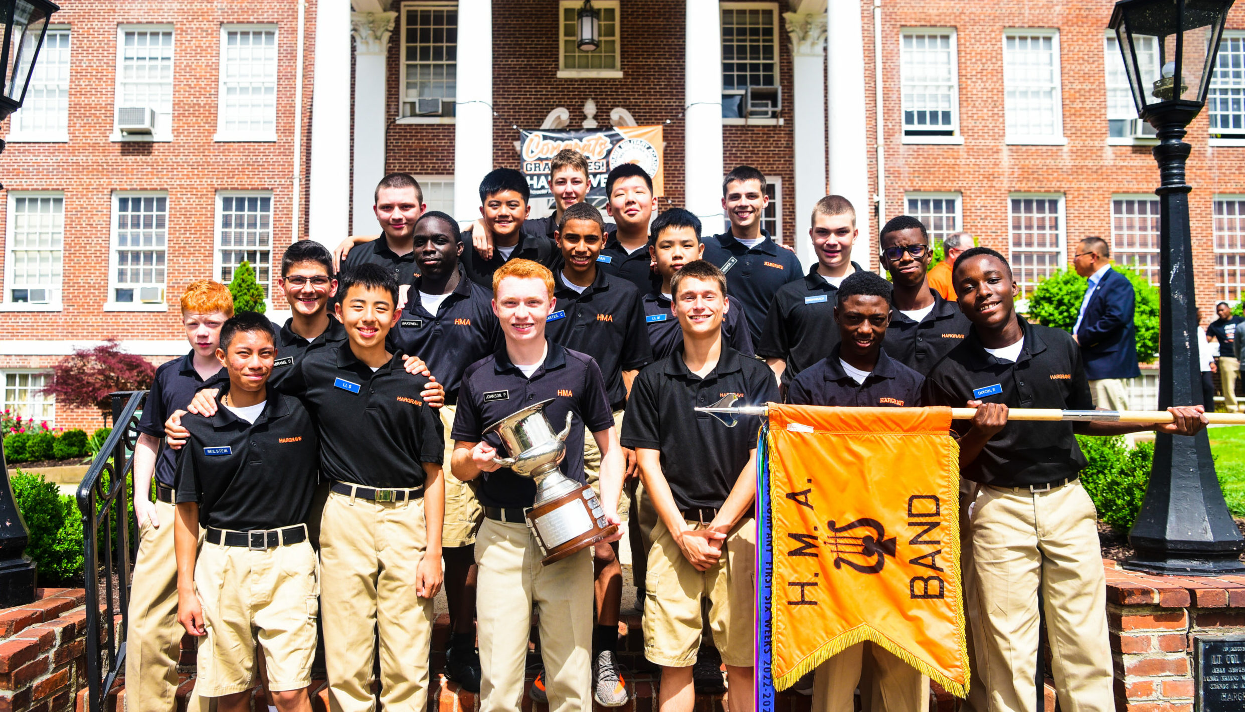 Band Company cadets holding flag and trophy in front of main building.